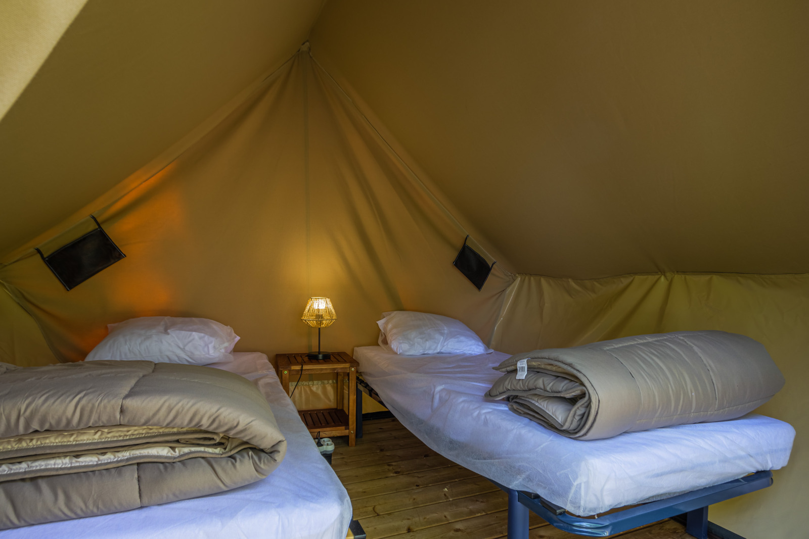 Binnenkant van een safaritent met twee eenpersoonsbedden, beddengoed, nachtkastje en lamp in het midden.