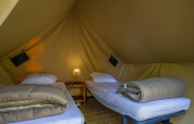 Interieur van een safaritent met twee eenpersoonsbedden, beddengoed, nachtkastje en een lamp tussenin.