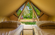 Vista interior de una tienda glamping con dos camas, edredones y entrada abierta hacia un camping verde.