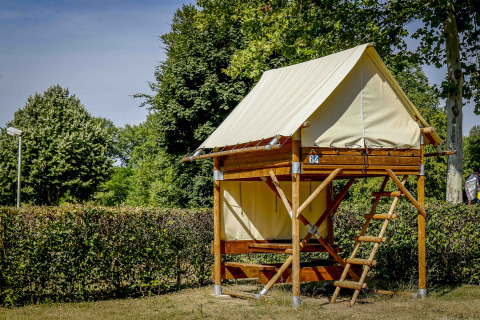 Tenda safari Bivouac Explorer su piattaforma rialzata in legno con scala, circondata da siepi e alberi verdi.