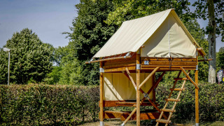 Tenda safari Bivouac Explorer su piattaforma rialzata in legno con scala, circondata da siepi e alberi verdi.