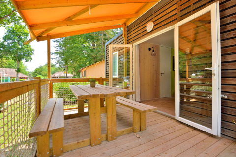 Terraza cubierta de madera con banco y mesa en Cottage New Valley, rodeada de árboles verdes.
