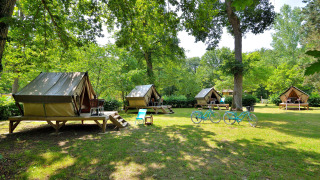 Photo of several teepee tents on a grassy campsite, two blue bikes, and outdoor chairs under green trees.