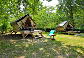 Two elevated teepee-style tents in a green forest, featuring decks and outdoor chairs for relaxation.