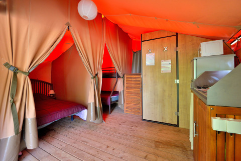 Interieur van de Lodge Victoria safaritent met houten vloer, bed, stapelbedden en keuken in Franse camping.