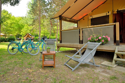 Zona de camping al aire libre con bicicletas azules, silla plegable y flores ante la tienda safari Lodge Victoria.