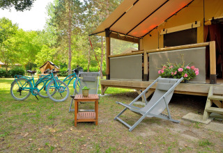 Zona de camping al aire libre con bicicletas azules, silla plegable y flores ante la tienda safari Lodge Victoria.