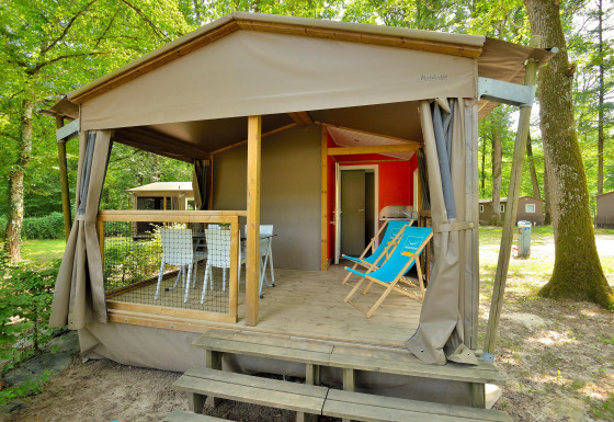 Photo of a Mobilodge cabin with a covered porch, deck chairs, dining set, and surrounded by forest trees.