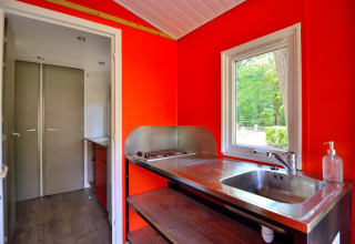 Innenansicht einer Mobilodge-Küche auf Camping Seasonova Etang de la Vallée mit roter Wand und Fenster.
