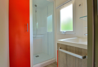Modernes Badezimmer mit roter Schranktür, Dusche, Fenster und Waschbecken in der Mobilodge auf Camping Seasonova Etang de la Vallée, Frankreich.