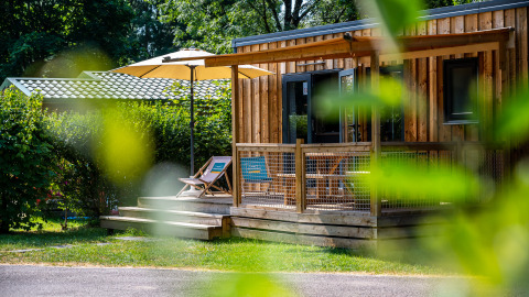 Glampingverblijf met houten chalet, terrasstoelen en parasol, omgeven door weelderig groen.