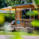 Glamping-ophold med træhytte, terrasse, liggestole og parasol omgivet af grønne planter og natur.