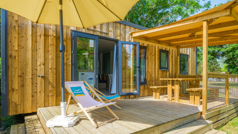 Moderne glampingverblijf met houten terras, ligzetel, parasol en overdekte picknickruimte buiten.