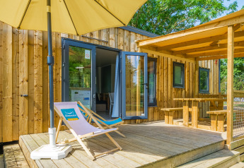 Moderne glampingverblijf met houten terras, ligzetel, parasol en overdekte picknickruimte buiten.
