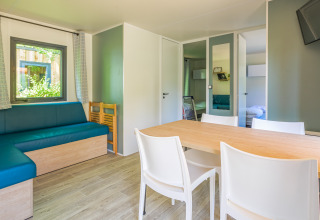Alojamiento glamping moderno y luminoso con mesa de comedor, sillas blancas, sofá azul y televisor.
