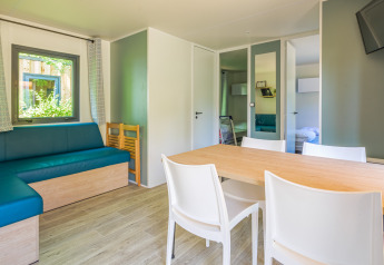Alojamiento glamping moderno y luminoso con mesa de comedor, sillas blancas, sofá azul y televisor.