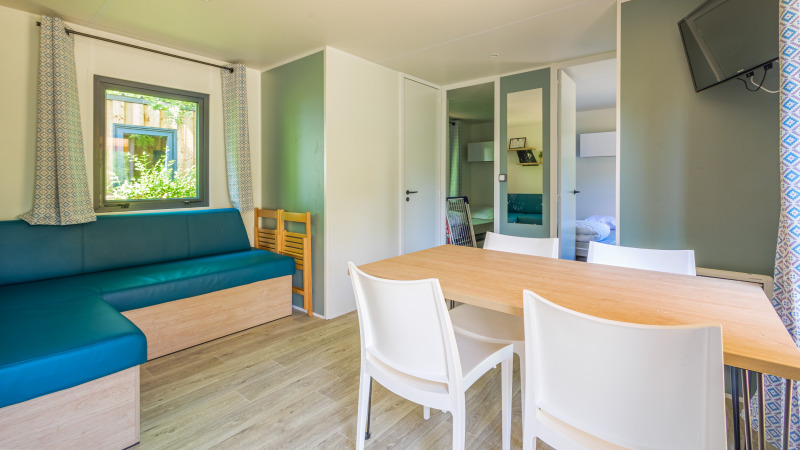 Alojamiento glamping moderno y luminoso con mesa de comedor, sillas blancas, sofá azul y televisor.