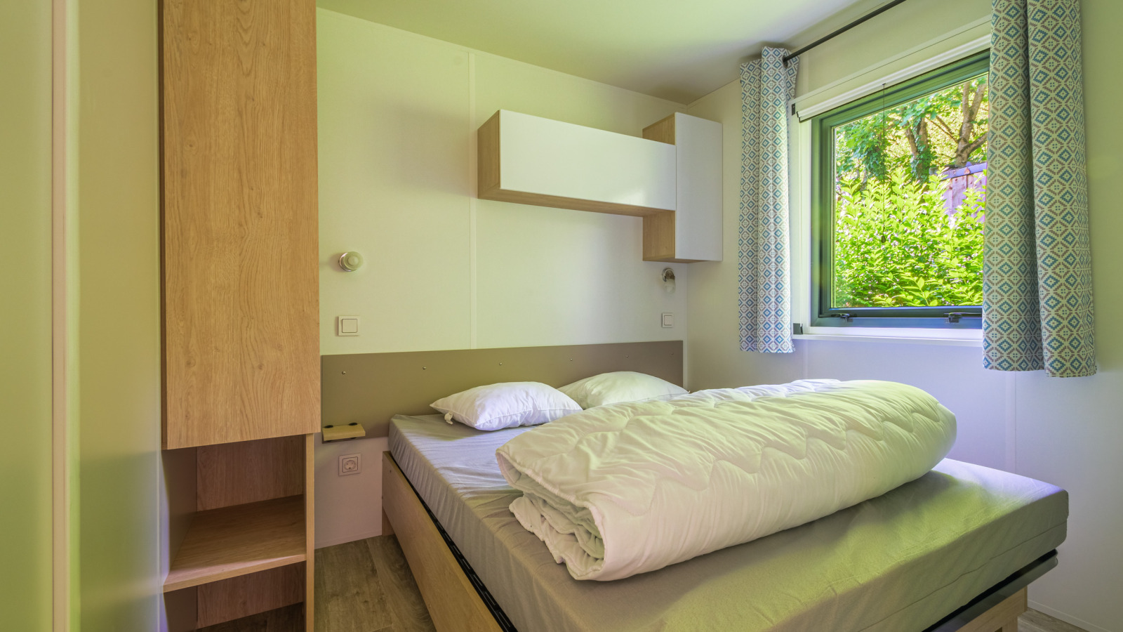 Lichte glampingkamer met tweepersoonsbed, moderne houten kasten en raam met uitzicht op groen.