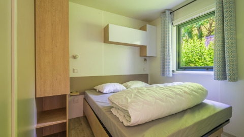 Lichte glampingkamer met een tweepersoonsbed, houten kasten en een raam met zicht op struiken.