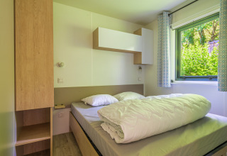 Lichte glampingkamer met een tweepersoonsbed, houten kasten en een raam met zicht op struiken.