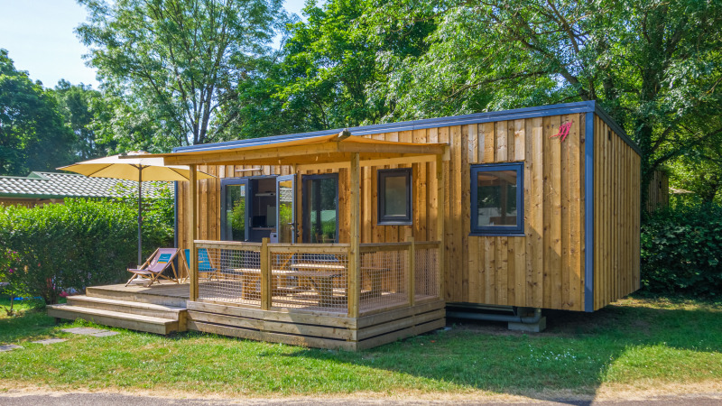 Alojamiento glamping moderno de madera con terraza, sombrilla y tumbonas en un entorno natural verde.