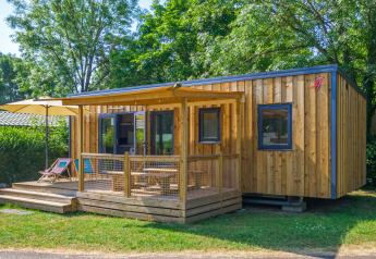 Alojamiento glamping moderno de madera con terraza, sombrilla y tumbonas en un entorno natural verde.