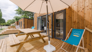 Terrazza esterna in legno con tavolo da picnic, ombrellone e sdraio davanti a una cabina glamping moderna.