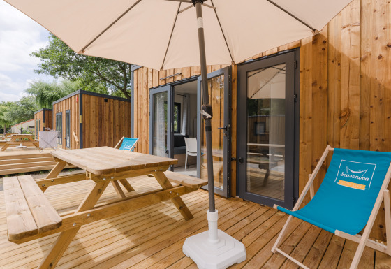 Outdoor wooden terrace with picnic table, sun umbrella and deckchair in front of a modern glamping cabin.