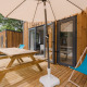 Udendørs træterrasse med picnicbord, parasol og liggestol foran moderne glamping-hytte i naturen.