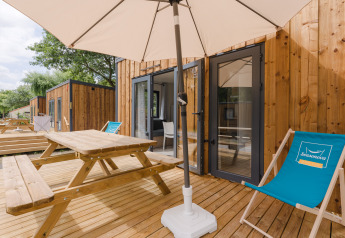 Udendørs træterrasse med picnicbord, parasol og liggestol foran moderne glamping-hytte i naturen.