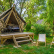 Glamping tipi telt på en træplatform i en grøn skov, med terrasse, borde, stole og solstole udenfor.