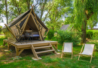 Modern teepee tent on a wooden platform in a lush forest, with patio table, chairs, and two loungers.