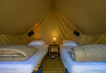 Inside a teepee tent with two single beds, bedding, and a small lamp on a nightstand between them.