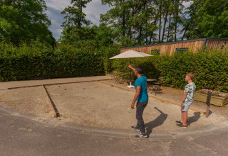 To personer spiller petanque på en grusbane ved Camping Seasonova Vittel, omgivet af grønne buske.