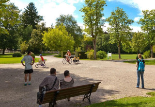 Folk nyder det solrige vejr i en grøn park ved Camping Seasonova Vittel i Grand Est, Frankrig, med cyklister og bænke.