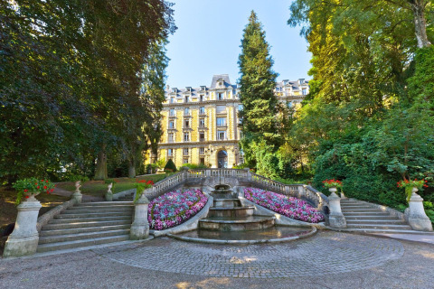 Elegantes Herrenhaus im Camping Seasonova Vittel, eingerahmt von Treppen, Blumen und Bäumen in Grand Est, Frankreich.