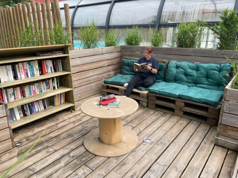 Outdoor reading nook with bookshelf, pallet bench, green cushions, and a person reading on a wooden deck.