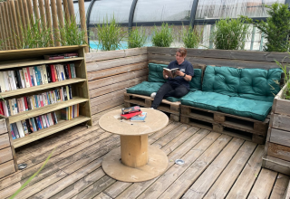 Outdoor reading nook with bookshelf, pallet bench, green cushions, and a person reading on a wooden deck.