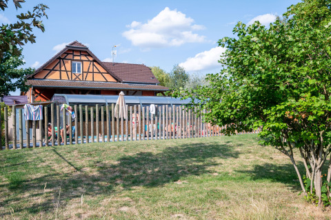 Holiday park with half-timbered house, fenced swimming pool and guests on a sunny day in Grand Est, France.