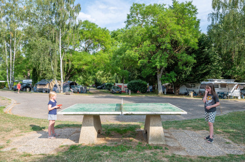 Two children play table tennis outdoors at a campsite with tents and caravans among lush green trees.
