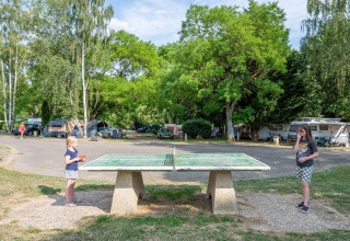 Two children play table tennis outdoors at a campsite with tents and caravans among lush green trees.