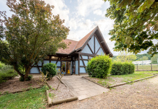 Hus med saddeltag og træramme omgivet af grønne buske ved Camping Seasonova Les Portes d'Alsace i Frankrig.