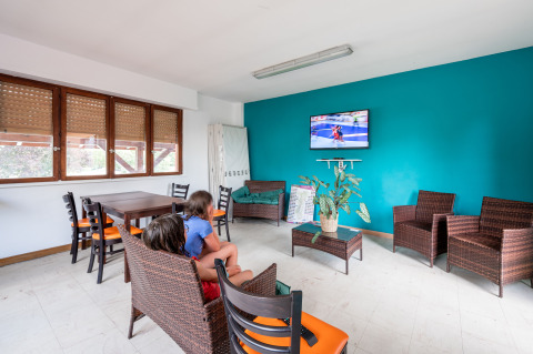 Bright lounge area at Camping Seasonova Les Portes d'Alsace with wicker chairs, kids watching TV, and a teal accent wall.