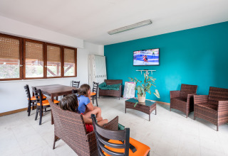 Bright lounge area at Camping Seasonova Les Portes d'Alsace with wicker chairs, kids watching TV, and a teal accent wall.