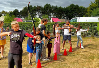 Niños y adultos practican tiro con arco en una zona verde junto a tiendas safari en Camping Seasonova Etennemare.