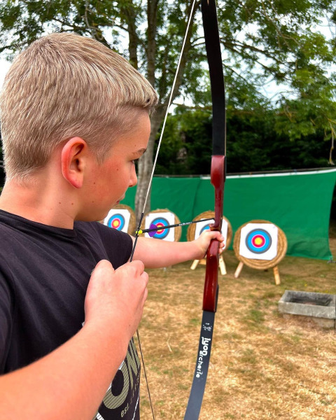 Niño rubio apunta con arco y flecha a dianas en Camping Seasonova Etennemare, parque vacacional en Normandía, Francia.