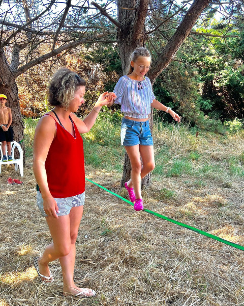 Woman helps girl balance on a slackline among trees at Camping Seasonova Etennemare in Normandy, France.