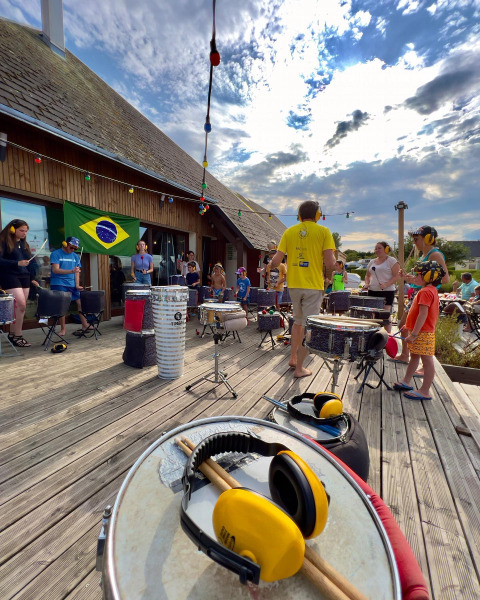 Taller de percusión al aire libre en Camping Seasonova Etennemare, con niños y adultos bajo bandera brasileña.