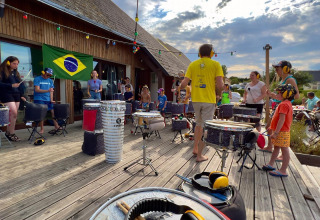 Outdoor drumming workshop at Camping Seasonova Etennemare with children and adults under a Brazilian flag.