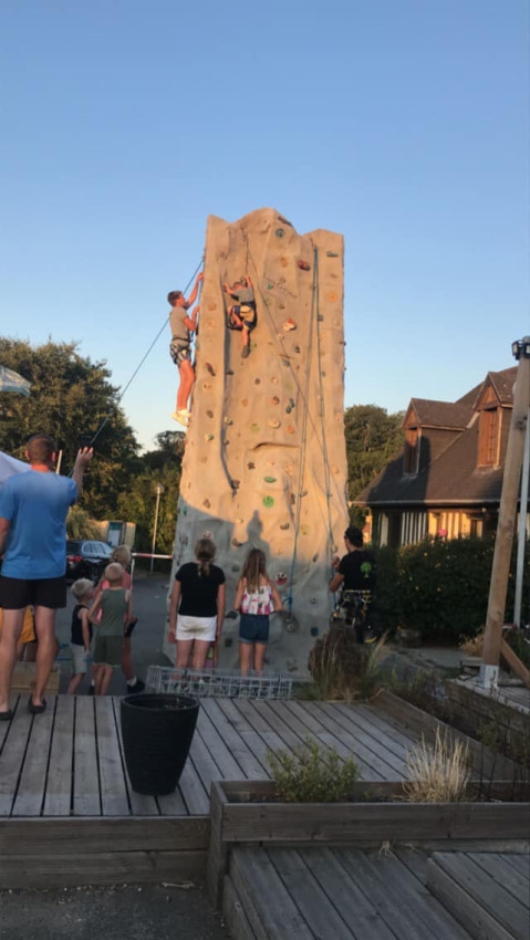 Menschen beobachten zwei Kletterer an einer Kletterwand auf einem sonnigen Campingplatz in der Normandie.